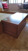 Full view of rectangular solid wood chest with dovetail side construction, medium brown finish, sitting on parquet floor near rocking chair and couch.