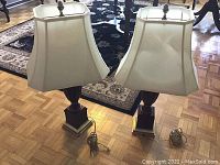Pair of vintage-style table lamps with white fabric shades shown on wood floor with area rugs in background.