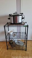 Photo of black metal wire utility shelf holding kitchen items such as a slow cooker on top and smaller kitchen utensils on lower shelves.