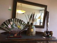Large rustic urn-shaped vase and Oriental folding fan on wooden stand on dark wood furniture under large mirror.