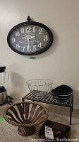 Photo showing the oval French inspired wall clock hanging on the wall above a metal stand holding three items: a wire basket, a small black lined basket, and a rusted metal decorative plate or bowl.