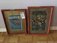 Two framed Japanese woodblock prints in red bamboo-style frames, one depicting a battle scene and the other a harbor scene.