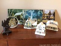 Full view of five ceramic Christmas village buildings and two decorated plates on wood surface.