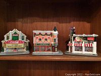 Front view of three Coca-Cola themed ceramic village figurines displayed on wooden shelf