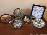 Group photo of six nature-themed collector plates on table, showing details of birds, animals and forest scenes, with one framed photo plate at back.