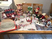 Overview of multiple Christmas themed items arranged on table including large pig figurine, various Santa and elf figures, glass plates, and a plaid textile.