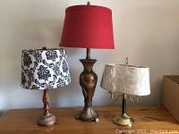 Three table lamps displayed on wooden surface, showing different sizes and styles including a large lamp with red shade, a medium lamp with damask shade, and small lamp with plastic-covered white shade.