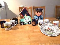 Photo of entire lot showing police police officer head figurine, ceramic police car coin bank, eight police-themed mugs, flag, and two decorative plates.