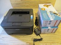 Brother black laser printer along with two cartridge boxes and cables on a surface.