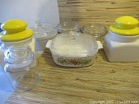 Overview photo showing multiple white glass casserole dishes with clear glass lids and two white square jars with bright yellow lids on a wood surface.
