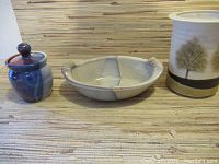 Three ceramic dishes shown side-by-side on a woven surface: a small covered jar, a divided shallow dish with two hand grips, and a cylindrical canister with a painted tree scene.