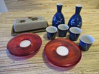 Full grouping shot showing all items: two ceramic bottles, four matching cups, two red glass candle holders, and a butter dish with floral lid on a striped surface.