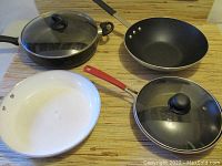 Four frying pans displayed on a wooden surface, two with glass lids, one black and one white ceramic coated, and another with a red handle.