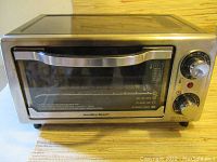 Front view of Hamilton Beach toaster oven showing stainless steel body, clear glass door with handle, and two control knobs for temperature and timer.
