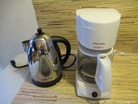 Electric kettle and Black & Decker VersaBrew coffee maker together on a surface showing the kettle's reflective finish, black handle and lid knob, and the coffee maker's white body and glass carafe with handle.