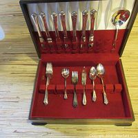 Cutlery set arranged inside wooden case, showing knives in lid and spoons/forks in bottom portion with red velvet lining.