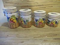 Front view of four ceramic storage jars of varying sizes arranged in a row with colorful fruit painted designs and woven basket bottom pattern on a wooden surface.