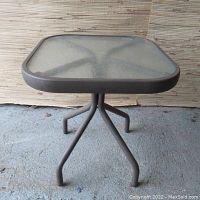 Full view of vintage outdoor side table showing square glass top and three metal legs supporting it.