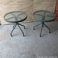 Pair of green metal framed outdoor side tables with round textured glass tops set on concrete floor.