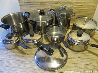 Photo shows nine stainless steel pots and pans with lids in varying sizes, some with black plastic handles, arranged on a surface.