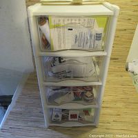 Front view of the white plastic four-drawer storage shelf filled with arts and craft materials including papers, fabric pieces, and other items.