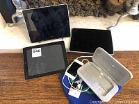 Overview of the lot showing three black Apple iPads, two white Apple iPods, and charging cables on a wooden surface.
