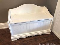 White wooden blanket box with paneled front, curved backrest lid and scalloped base trim against wall on dark wood floor.