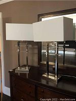 Pair of chrome and acrylic table lamps side by side on dark wooden dresser in front of mirror.