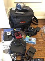 Lot showing cameras, camera bag, remotes, chargers and cables arranged on floor