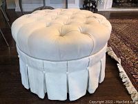 Front side view of the round beige ottoman with button-tuck top and pleated skirt base, showing fabric condition and details.
