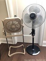 Two electric floor fans side by side; one beige Holmes pedestal blower fan on stand and one black Holmes oscillating pedestal fan.