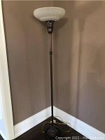 Full view of the tall floor lamp standing against a wall showing the base, stand, and glass shade at the top.