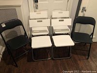 Six folding chairs shown folded and standing; four white and two black.