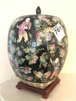 Front side of the Oriental ginger jar showing intricate multi-colored floral patterns and children figures on a black background with wooden base.