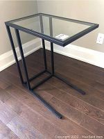 Side angle view of the glass and steel side table against a wall showing the cantilever design and black steel frame.