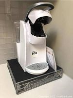 Side view of white Keurig coffee maker on top of metal mesh drawer stand displaying open lid and drip tray.