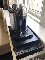 Nespresso VertuoLine coffee machine sitting on black metal drawer stand with sliding mesh drawer for coffee pods.