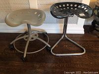Image showing both tractor seat stools side by side, beige stool with five caster wheels and black stool with chrome triangular base.
