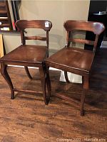 Front and side view showing pair of wooden billiard stools with curved legs and backrest, dark finish, placed on wood floor.