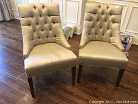 Front view of two matching beige button tuck upholstered chairs with dark wooden legs on brown floor in a room with panelled walls.