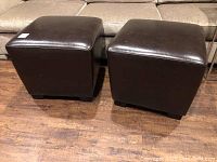 Two cube ottomans placed side by side on a wooden floor in front of a beige sofa. Dark brown leather or faux leather upholstery visible with cushioned tops.