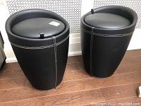 Pair of black storage stools side by side on a wooden floor near a white patterned wall. Both stools have a barrel shape, black faux leather upholstery with white stitching, and cushioned tops with tabs for lifting.