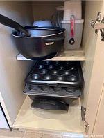 Inside cupboard showing a black pot with handle on top shelf and stacked muffin tins below.
