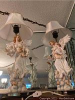 Pair of ceramic angel table lamps with shades on a reflective surface, showing full lamp with angel holding baby angel figures.