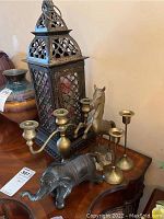 Photo shows brass candelabras, horse and elephant figurines, and metal glass lantern on wooden surface
