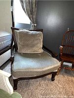 Front view of the dark wood wingback chair showing stained light quilted seat cushion and back pillow, curved wooden armrests and legs, on patterned rug in room.