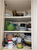 Three shelves filled with mixed dishware including plates, bowls, mugs, glass items and bakeware with various colors and styles.