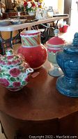Four ceramic items on a table: a floral teapot, two holiday themed pots, and a blue marbled pot.