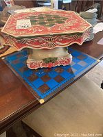 Photo of the ceramic cake platter on top, ornate red and cream decoration with floral and scroll motifs and pedestal base. Glass game board below with blue and brown checker pattern.
