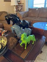 Three horse statues displayed on a wooden table. Black ceramic horse statue tall with visible paint loss, blue ceramic horse smaller, and green cast iron horse statue bright green in front.